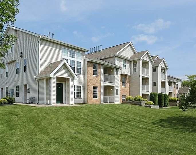 Charming multi-story apartment complex with a blend of brick and siding, surrounded by lush green lawns and mature trees under a clear blue sky.