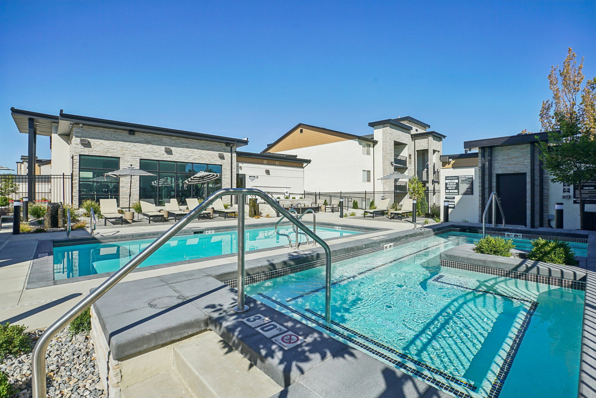 Luxurious apartment complex featuring a sparkling swimming pool, modern lounge chairs, and a stylish stone facade under a clear blue sky.