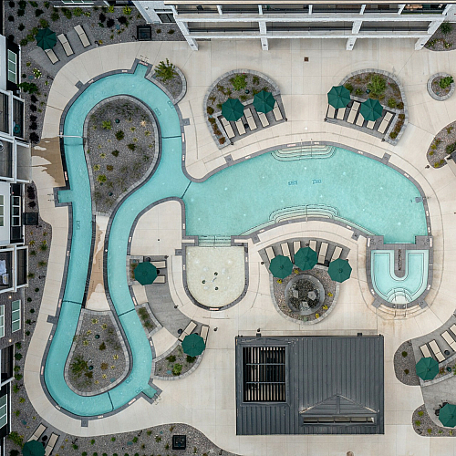 Aerial view of luxurious resort-style pool area with curving pools, spa, and numerous lounging spaces, surrounded by lush greenery and modern apartments.