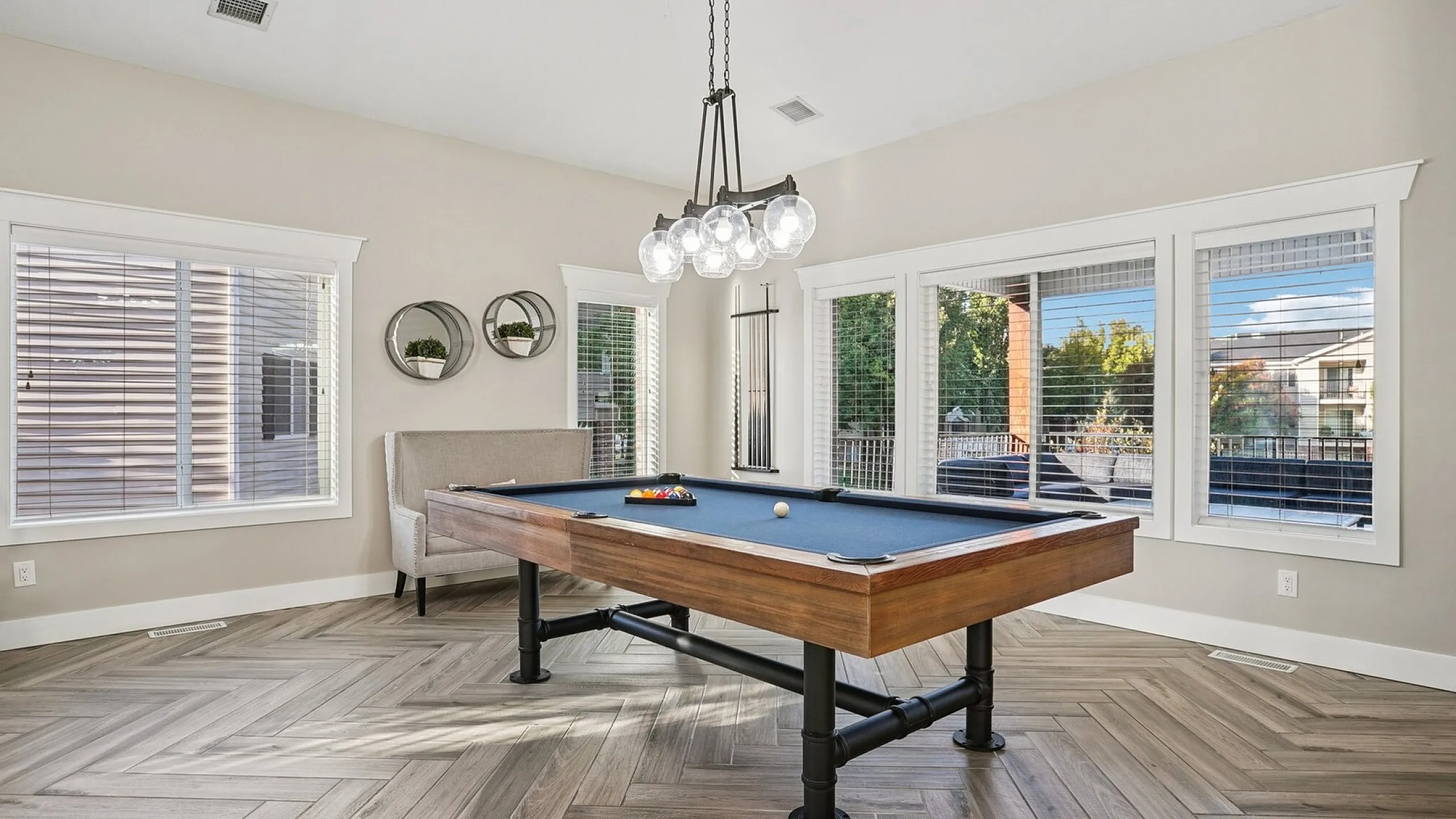 Chic game room with a sleek pool table, modern hanging lights, and elegant herringbone flooring, bathed in natural light from large windows.