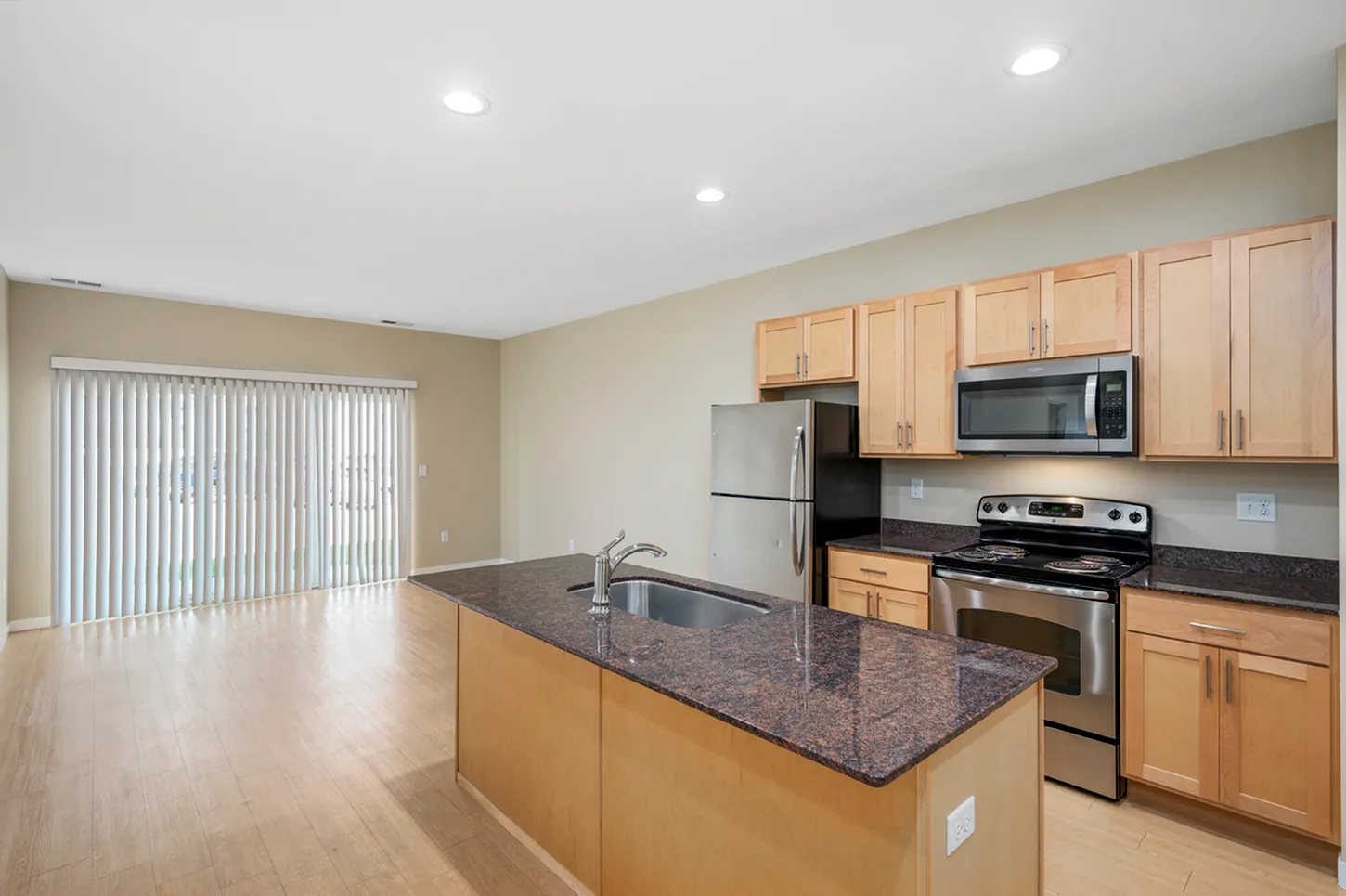 Modern open-concept kitchen with stainless steel appliances, elegant granite countertops, and light wood cabinetry opening to a spacious living area with natural light.