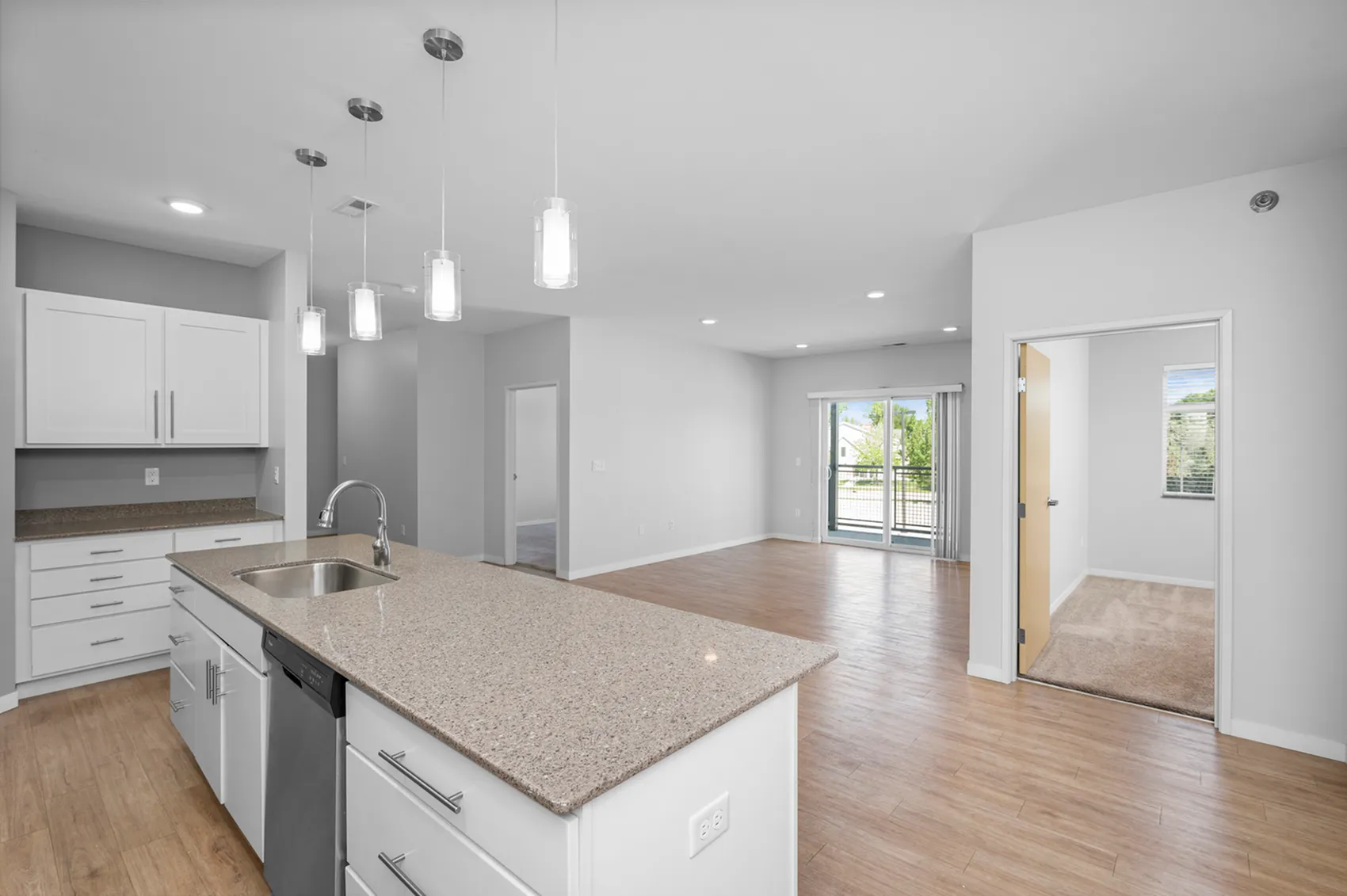Modern open-concept kitchen and living area with sleek white cabinets, elegant pendant lighting, and natural light from large windows opening to a spacious balcony.