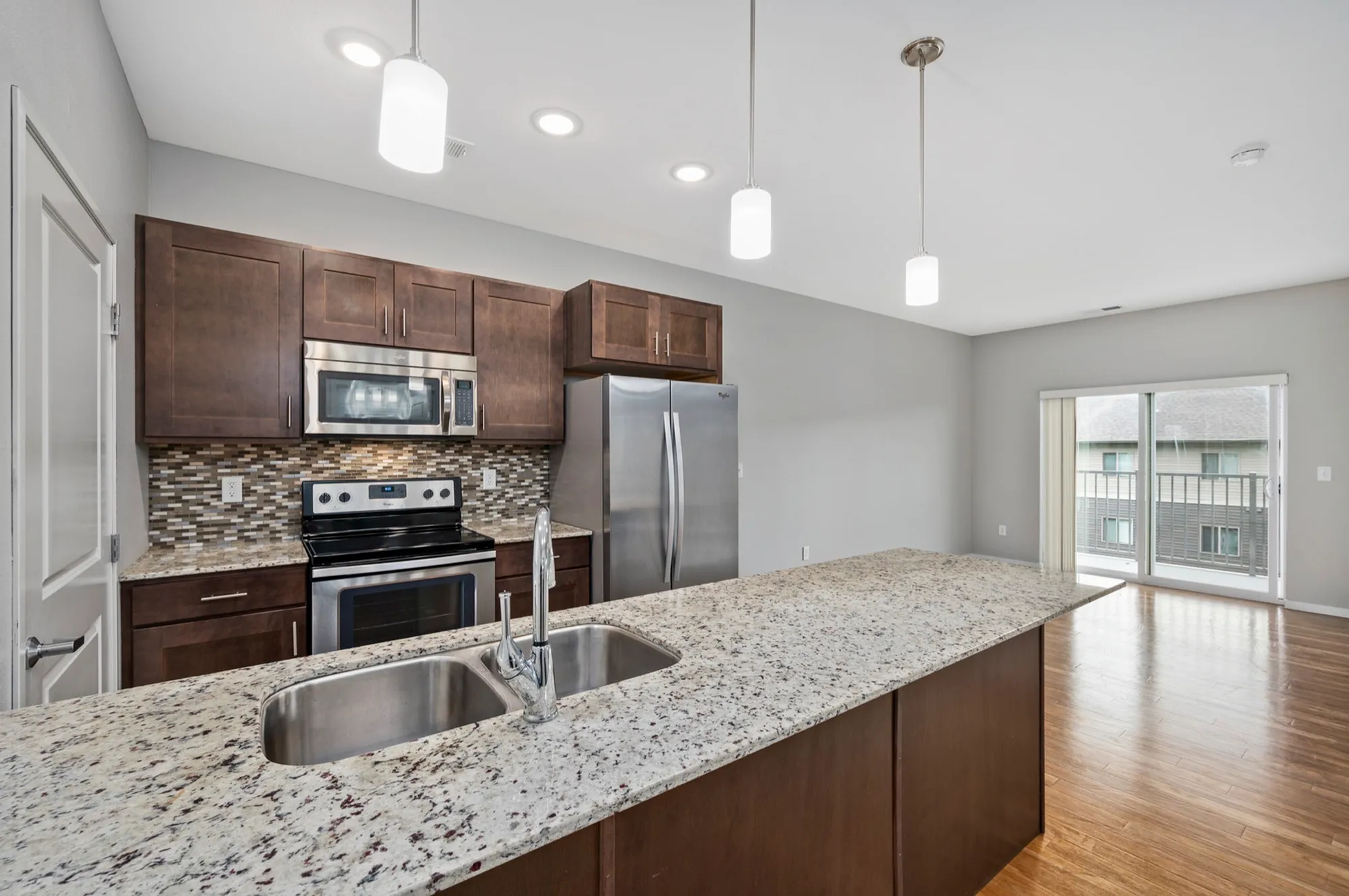 Modern kitchen with granite countertops, sleek stainless steel appliances, and elegant pendant lighting, opening to a bright living area with hardwood floors and a balcony view.