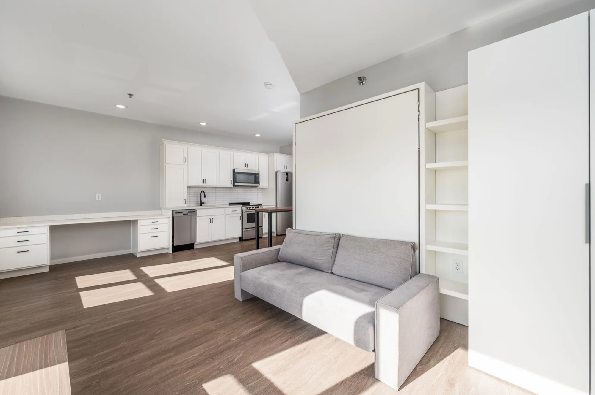 Modern open-concept living space featuring sleek kitchen with white cabinetry, integrated appliances, and a cozy gray sofa, enhanced by abundant natural light.