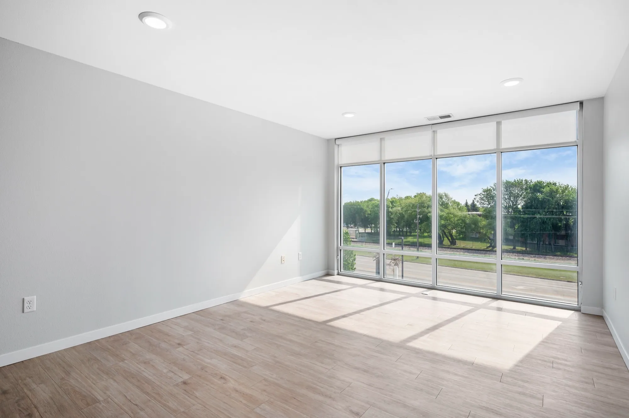 Bright and airy apartment room with large floor-to-ceiling windows offering a scenic view of greenery, complemented by sleek wood flooring and modern recessed lighting.