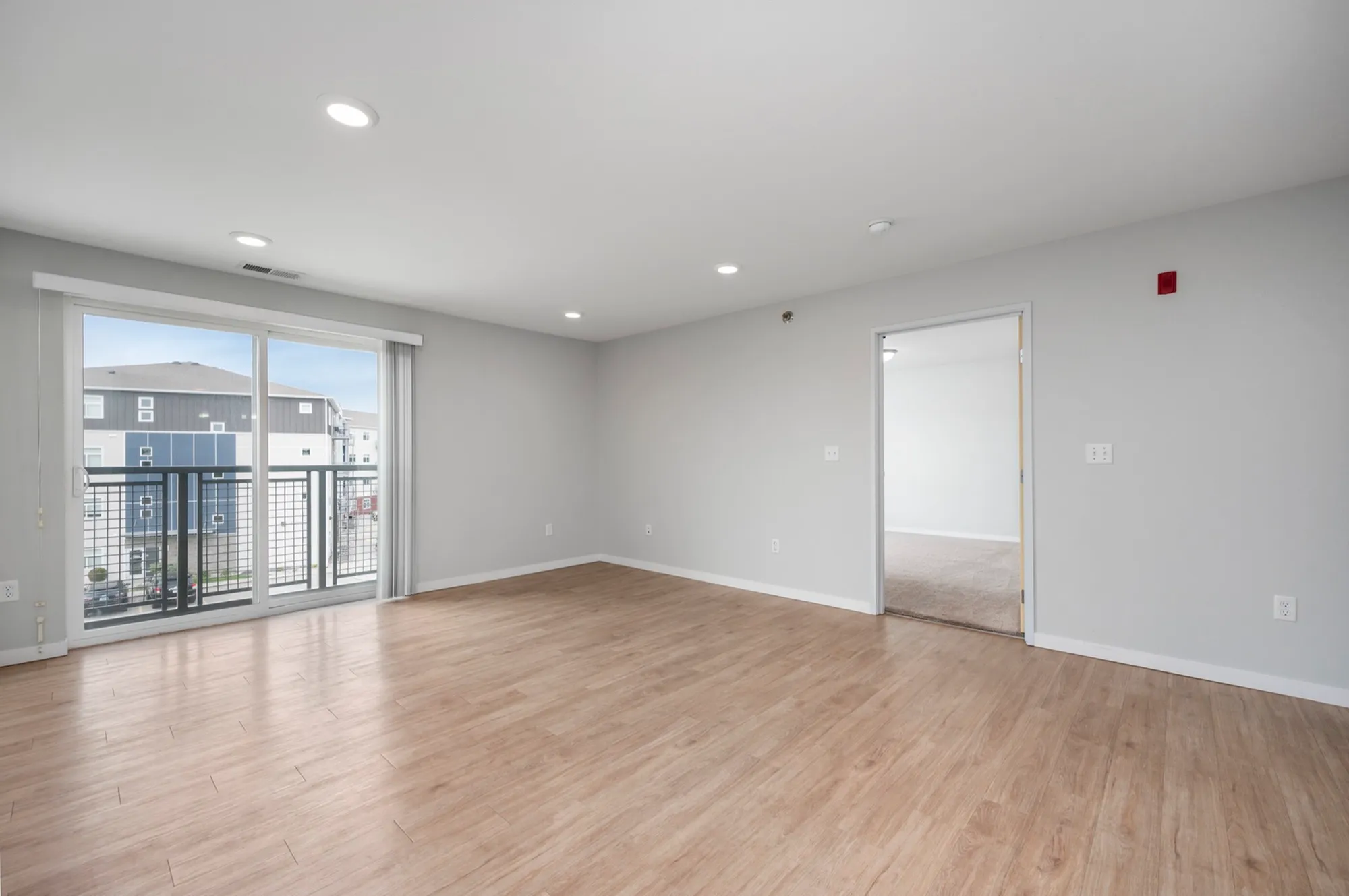 Bright and spacious living room with modern wooden flooring, large windows offering plenty of natural light and a view of the surrounding area, leading to a cozy adjacent room.