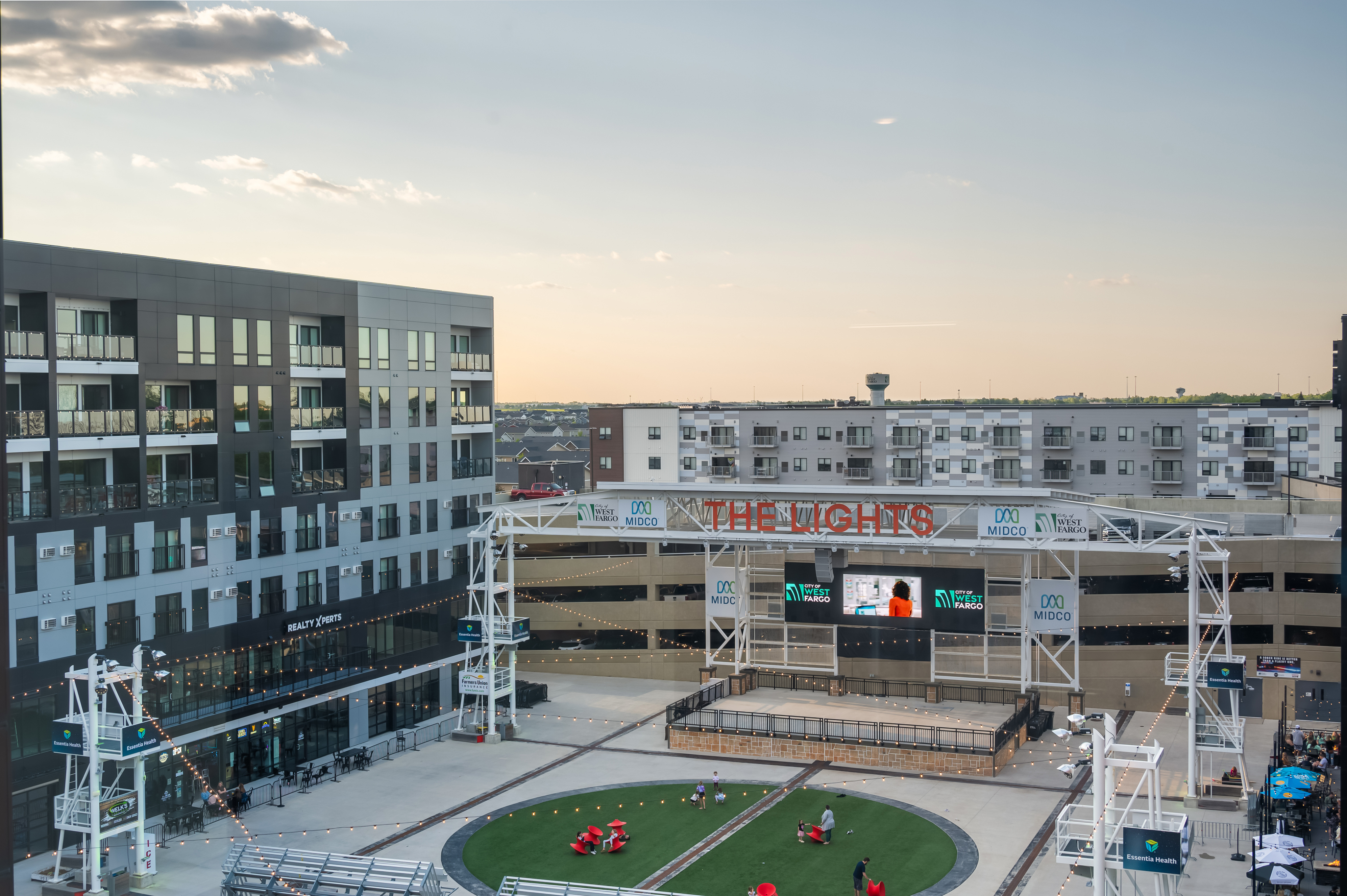 Vibrant urban complex with a modern residential building overlooking an inviting plaza and entertainment stage at sunset.
