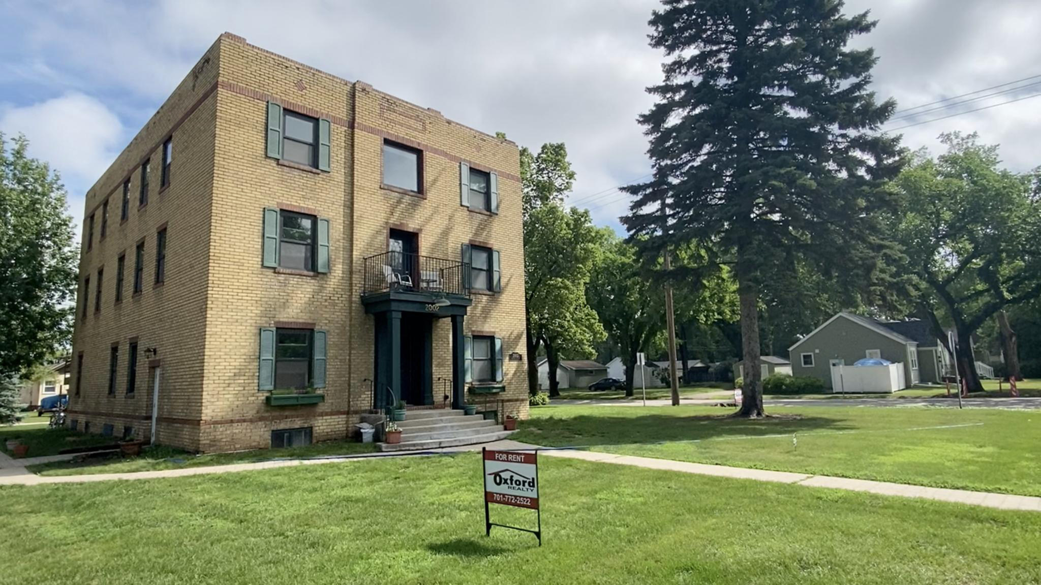 Charming three-story brick apartment building with classic architecture, lush green lawn, and cozy outdoor seating. Ideal location for vibrant living surrounded by tree-lined streets.