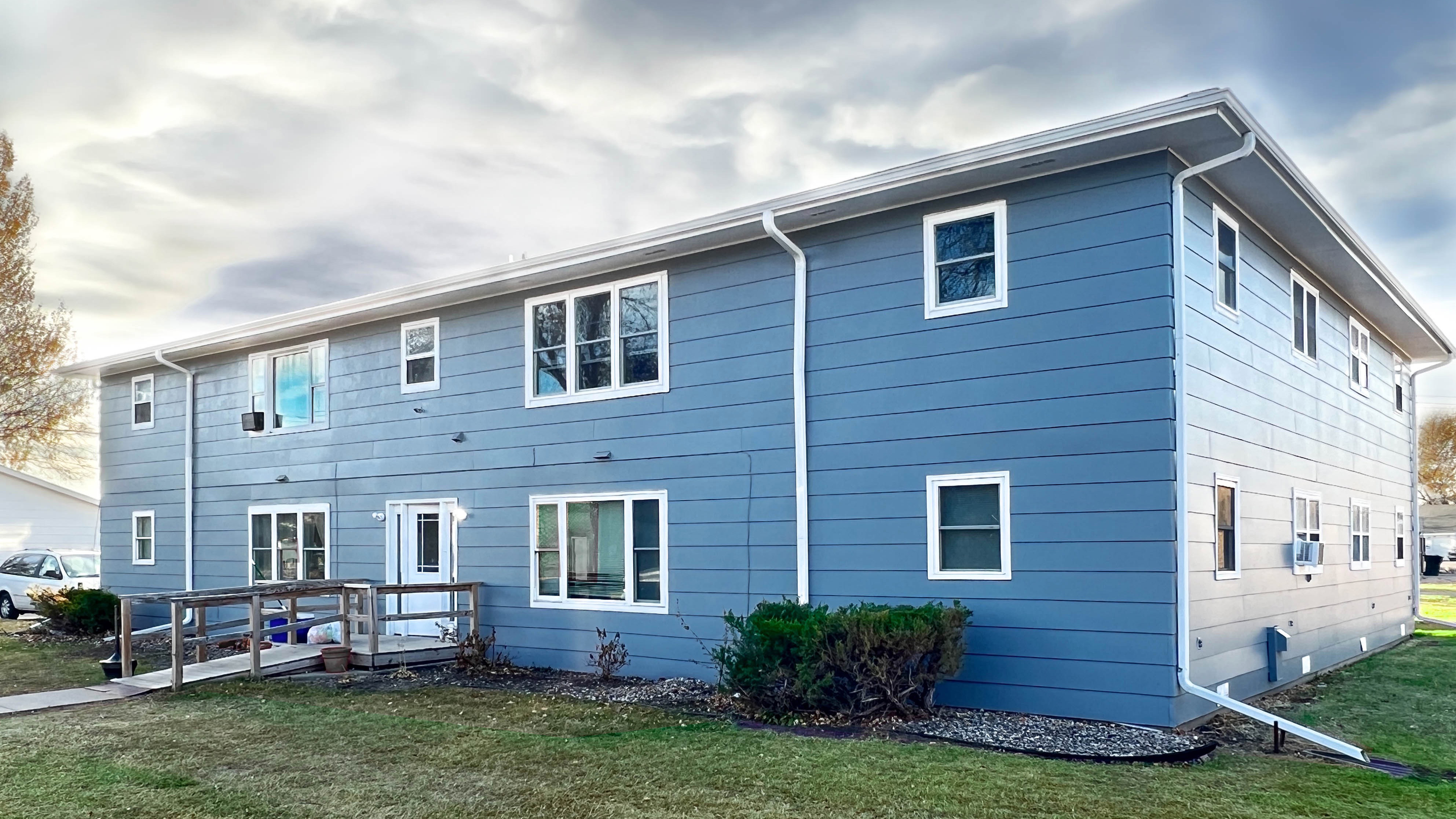 Charming two-story blue apartment building with symmetrical windows, surrounded by a lush green lawn and set against a cloudy sky.
