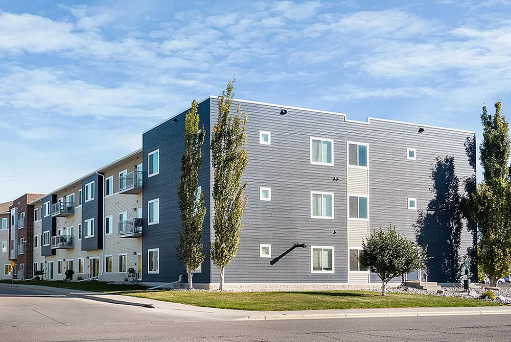 Modern apartment complex with sleek design, featuring multiple stories and stylish balconies, surrounded by lush greenery and a clear blue sky.