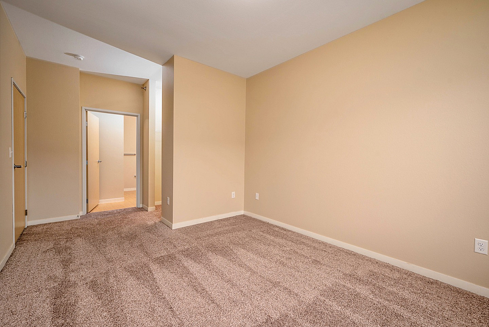 Spacious and well-lit empty room with clean carpeting, beige walls, and a door leading to an adjoining area.