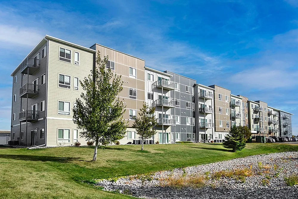 Modern four-story apartment building with private balconies, set in a lush landscaped area under a clear blue sky.