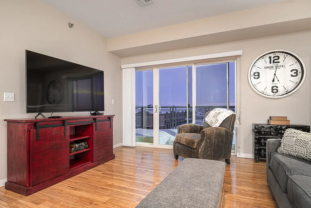 Chic living room with a large TV on a rustic red console, stylish seating, modern decor, and a wall clock, all accentuated by a bright balcony view.