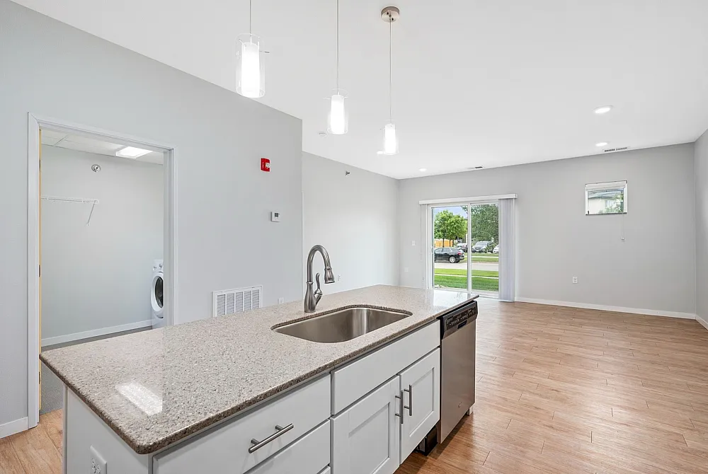 Bright modern kitchen with granite countertops, sleek cabinetry, dishwasher, and open layout overlooking a spacious living area with large windows and hardwood flooring.