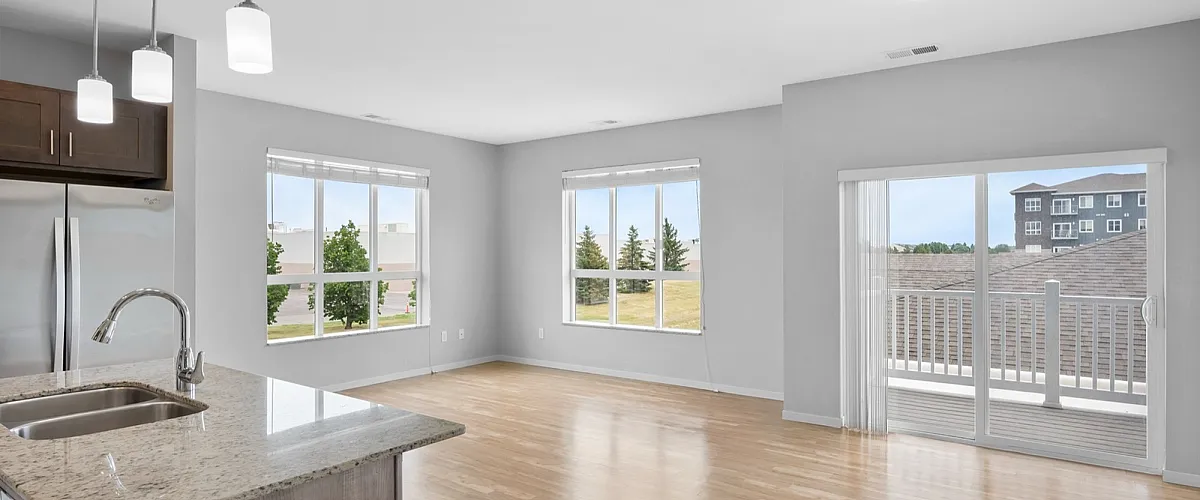 Bright and modern open-concept living space with elegant hardwood floors, a sleek kitchen island, and ample natural light from large windows and a sliding glass door leading to a balcony with serene views.