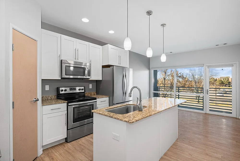 Modern kitchen with sleek stainless steel appliances, granite countertops, and stylish pendant lighting overlooking a bright living area with open balcony view.