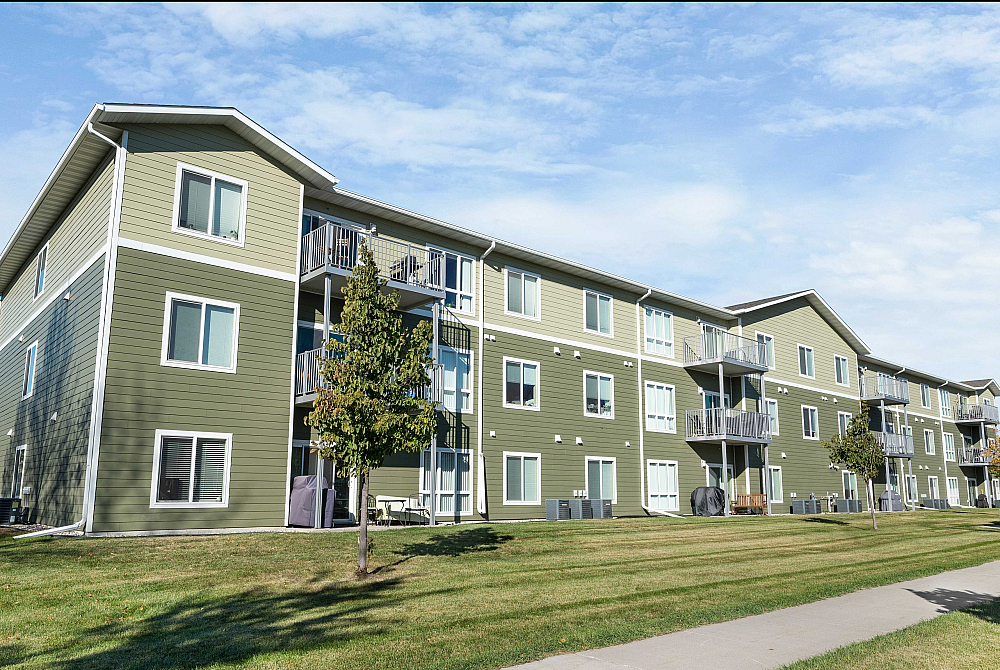 Charming three-story apartment building with modern green exterior and private balconies, surrounded by lush green lawns under a clear blue sky.