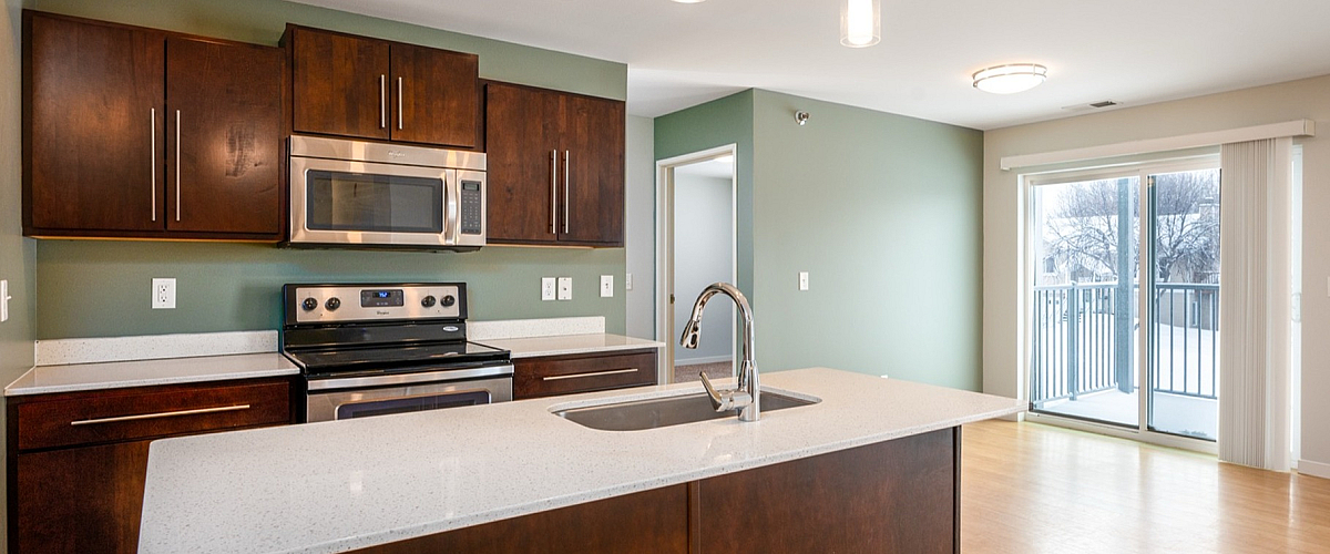 Modern kitchen with sleek wood cabinetry, stainless steel appliances, and a spacious island, overlooking a bright open living area with hardwood floors and large sliding doors to a balcony.