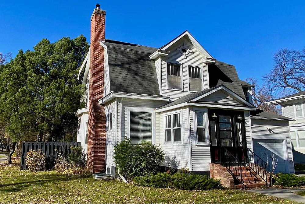 Charming two-story house with classic red brick chimney, nestled in a lush, tree-lined neighborhood under a clear blue sky.