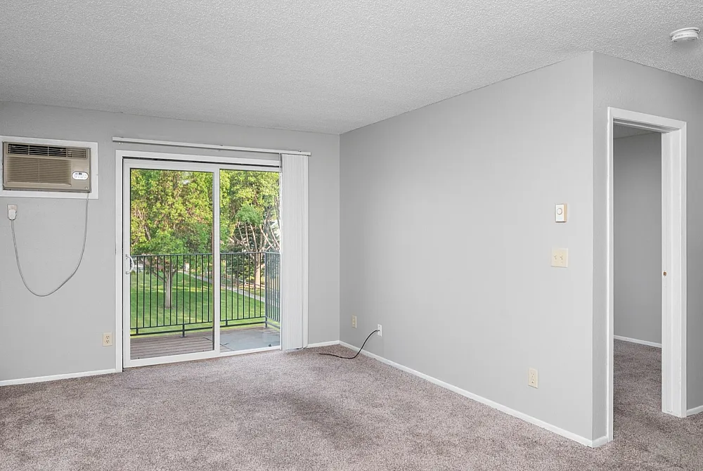 Bright living room with plush carpet, featuring a large sliding glass door leading to a balcony with lush green views.