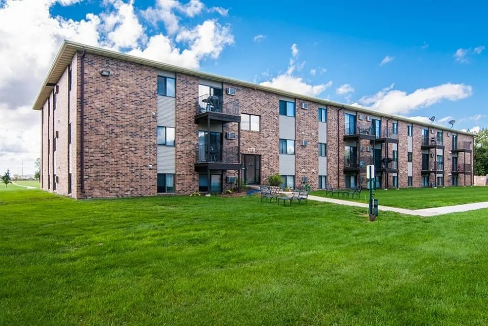 Charming three-story brick apartment building with balconies, set amid lush green lawns and a beautiful blue sky.