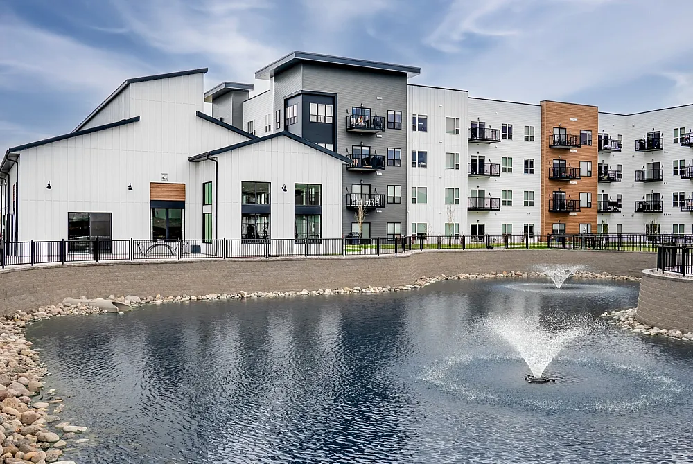 Modern apartment complex with sleek architecture, featuring balconies, large windows, and a serene pond with fountains in front.