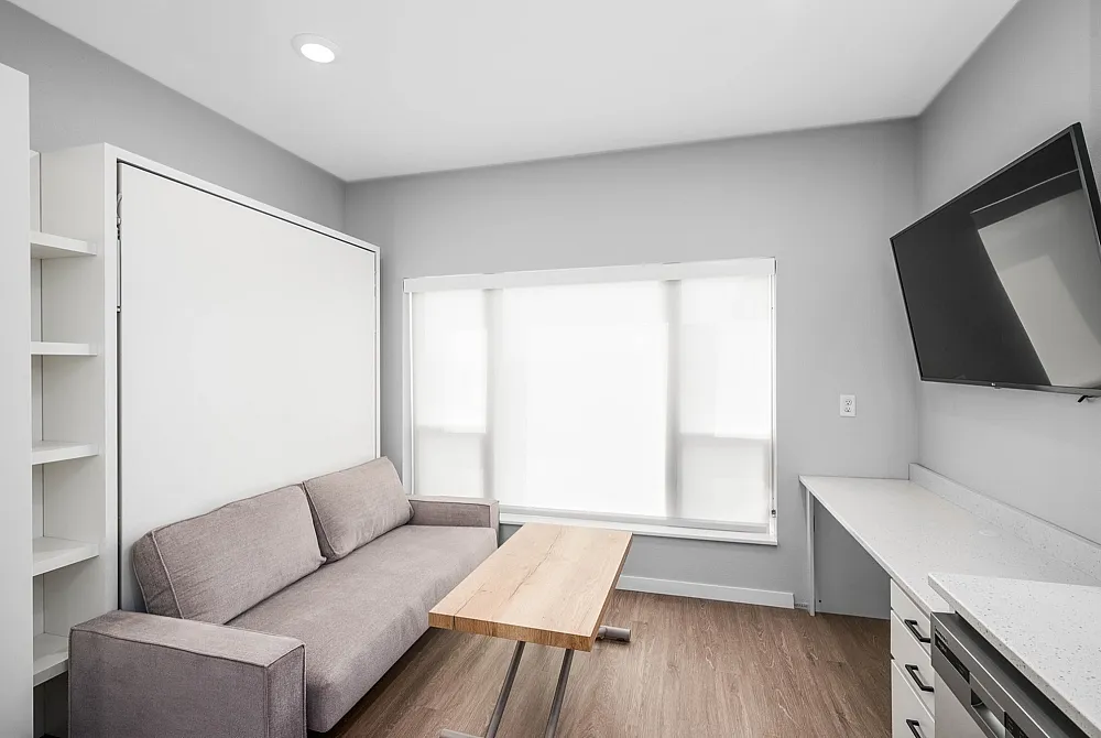 Modern apartment featuring a cozy gray sofa, sleek wooden coffee table, and wall-mounted TV, complemented by a bright, spacious design with a large window and elegant built-in shelving.