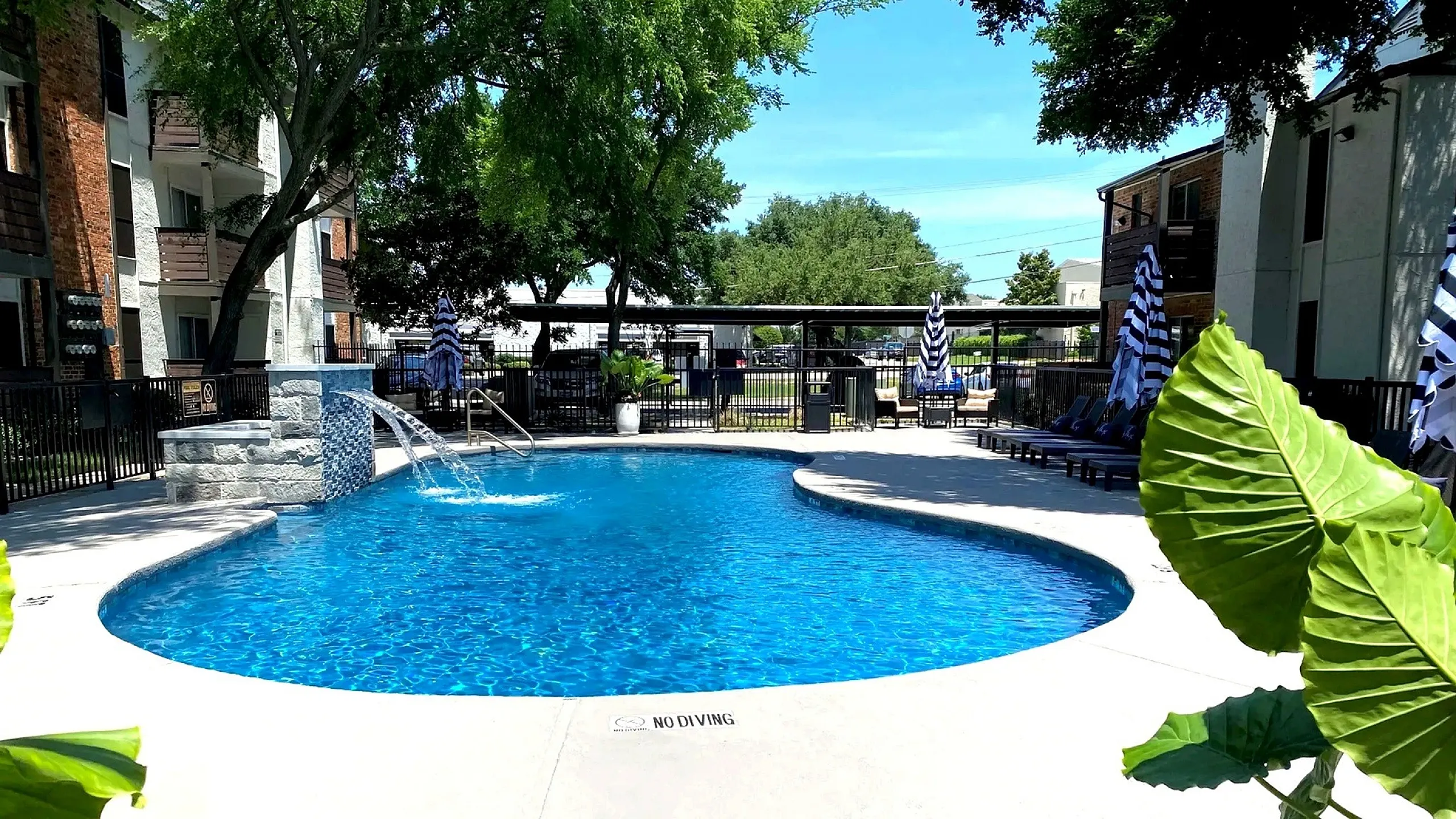 Inviting outdoor pool area with sparkling blue water and comfortable lounge chairs, surrounded by lush greenery and modern apartment buildings.