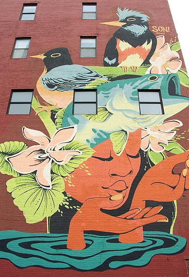 Vibrant mural on a brick building featuring colorful birds and a serene face surrounded by flowers and leaves, exuding creativity and urban charm.