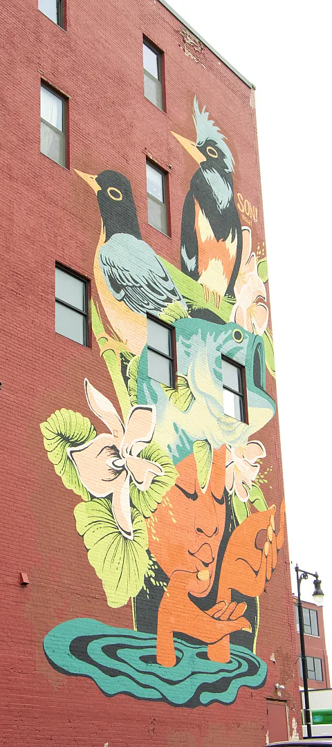 Vibrant street art mural on a brick building featuring colorful birds, lush greenery, and a serene face, adding artistic flair and cultural value to the urban scene.