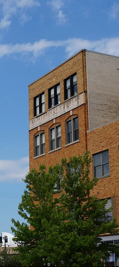 Charming urban apartment building with classic brick facade, framed by lush green trees against a bright blue sky.