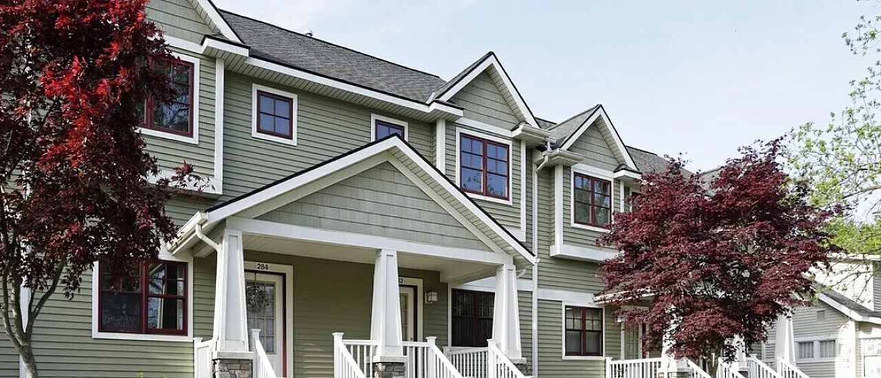 Charming two-story townhouse with green siding and red-trimmed windows, nestled in a beautifully landscaped garden with vibrant trees and shrubs.