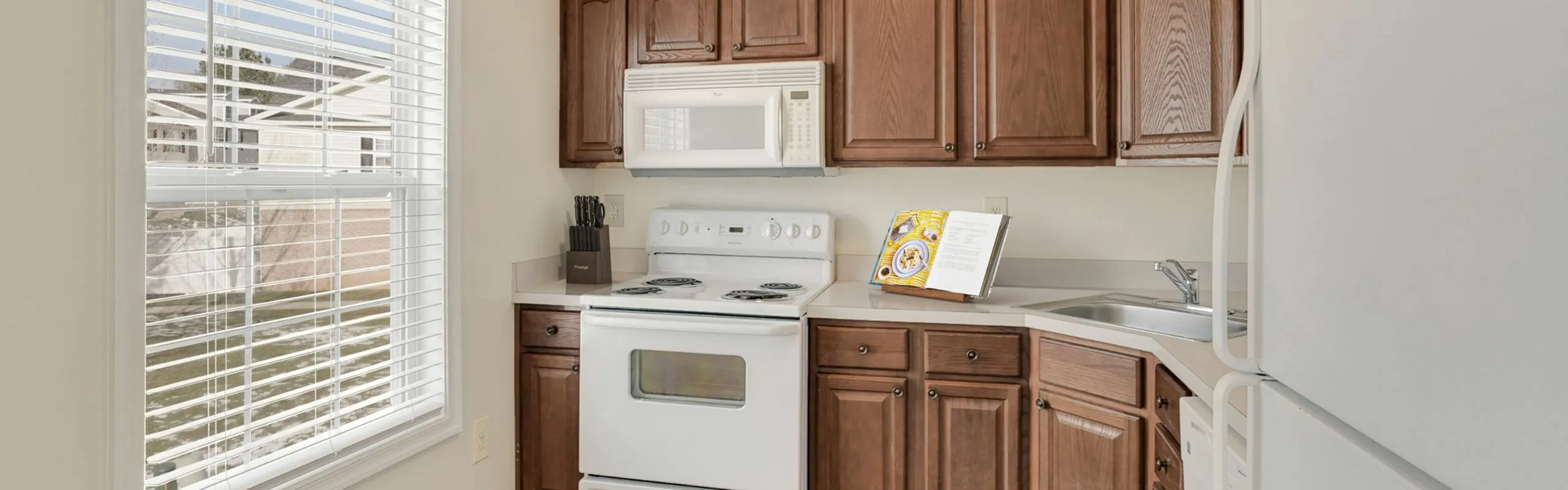 Bright kitchen with modern appliances, wooden cabinets, and large window with blinds, offering abundant natural light.