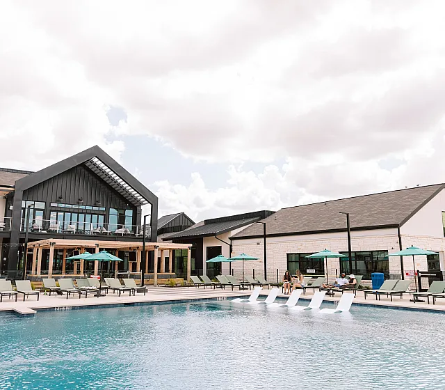 Elegant apartment complex showcasing a modern clubhouse, luxurious pool with lounge chairs, and inviting umbrellas under a bright sky.