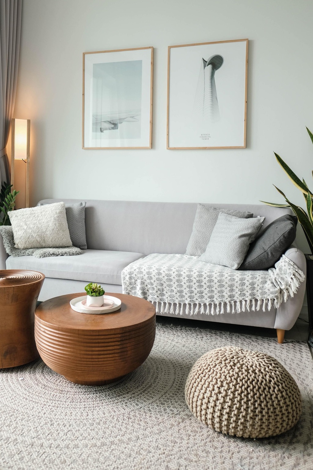grey couch with pillows. artwork on wall behind couch. 2 coffee tables and a small seat. plants on each side of the couch.