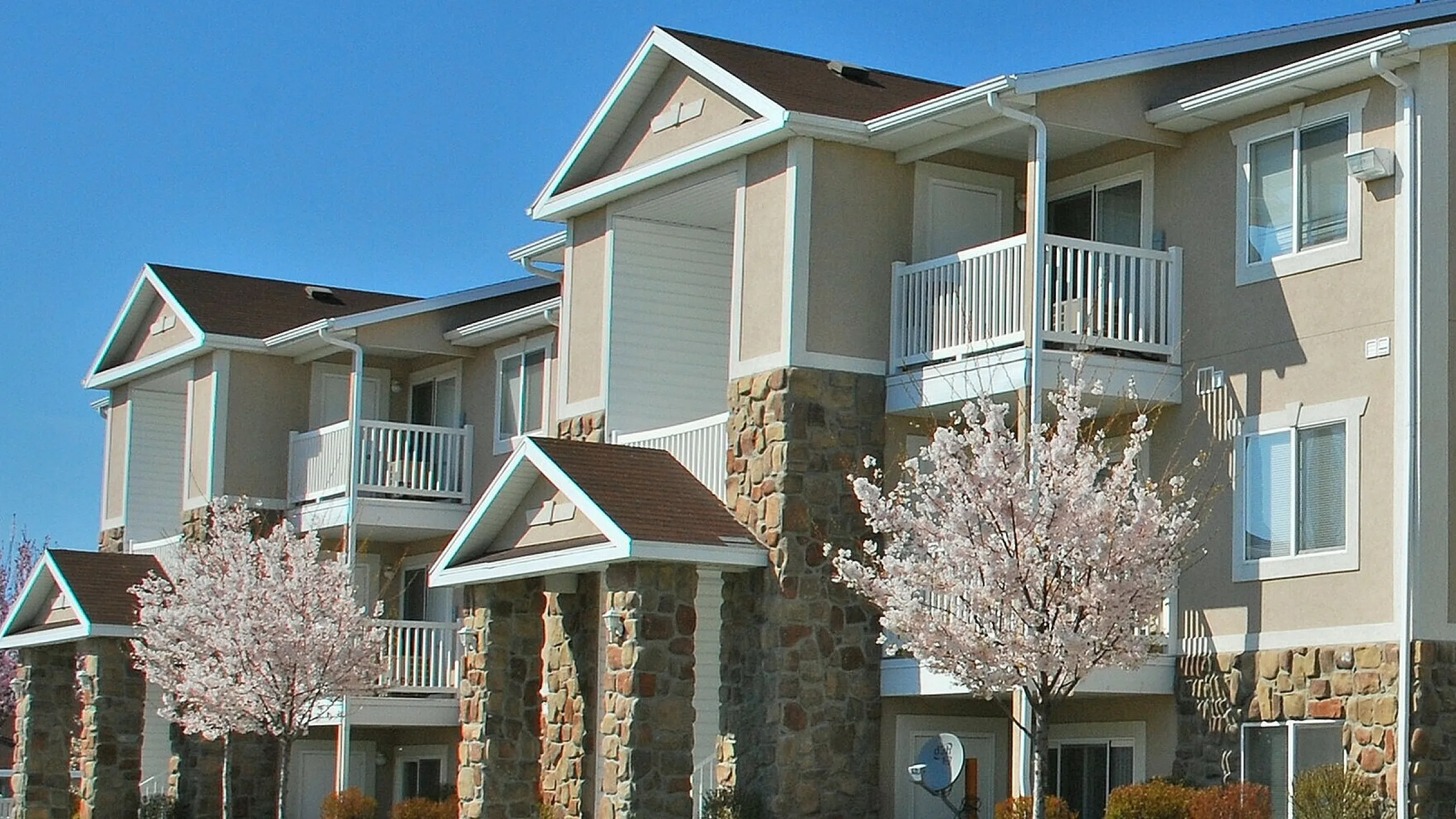 Charming apartment complex with stone accents and spacious balconies, surrounded by blooming cherry blossom trees and lush green lawns.