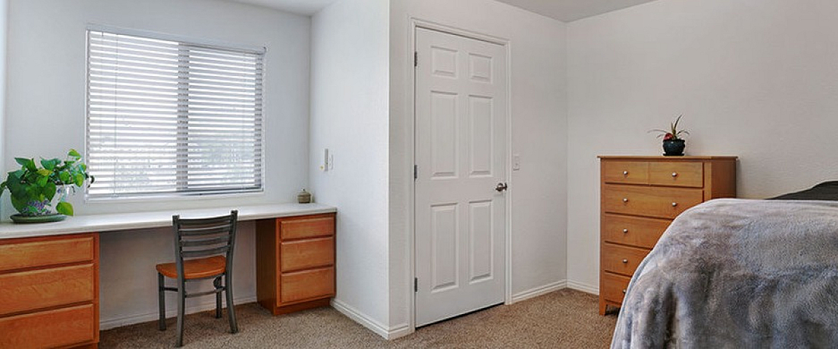 Bright bedroom with plush carpet, a cozy bed, elegant chest of drawers, and a well-lit desk area perfect for productivity.