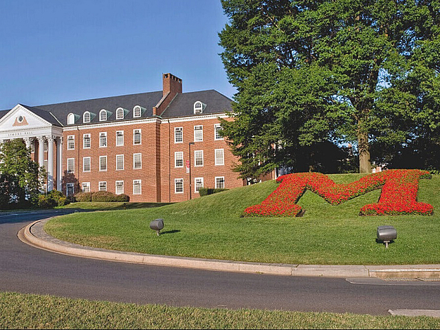 Elegant brick building with classic architecture surrounded by lush greenery and vibrant red flower shaped as the letter 'M', creating a welcoming atmosphere.