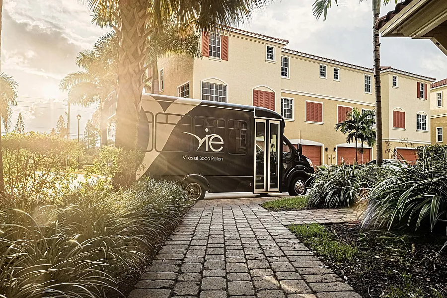 Sunny approach to luxurious Villas at Boca Raton featuring a modern shuttle service against a backdrop of elegant architecture and lush palm trees.