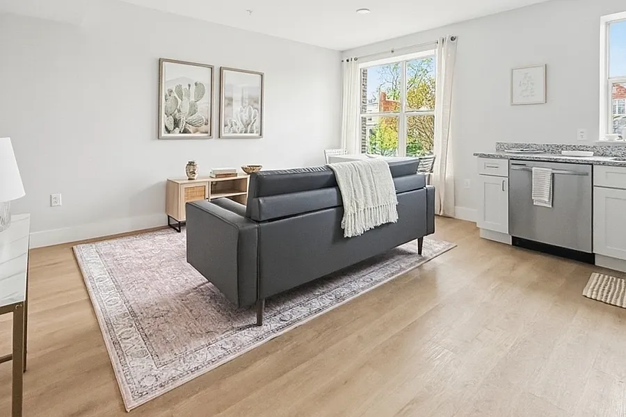 Bright and modern living space with a sleek grey sofa, stylish artwork, and large windows offering natural light. Open-concept design with a cozy kitchen area and elegant decor.