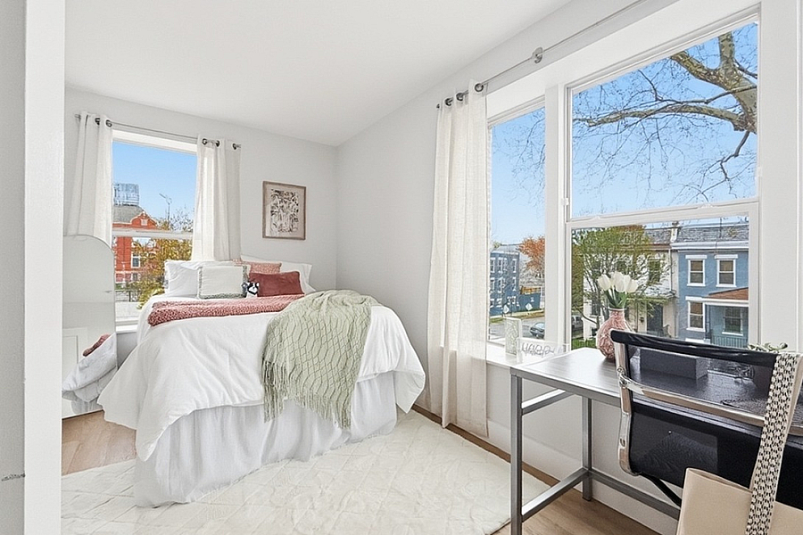 Bright and inviting bedroom with large windows offering picturesque neighborhood views, featuring a cozy bed and a modern workspace setup.