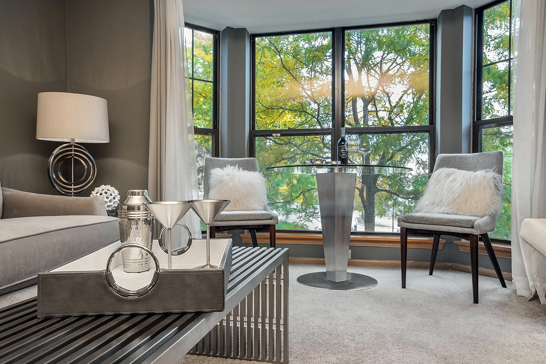 Chic living room with modern decor, large bay windows showcasing lush greenery, and cozy seating with plush cushions.