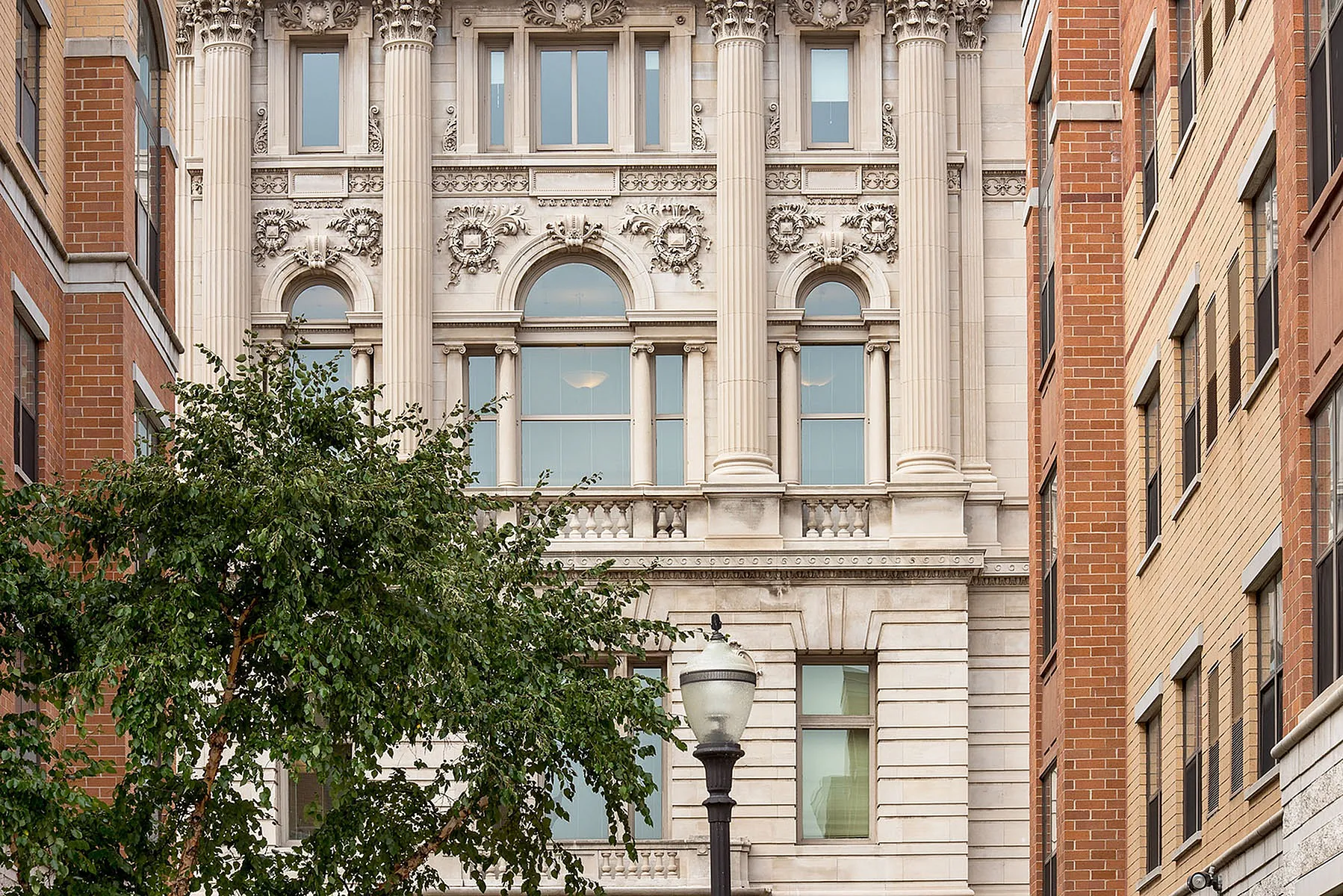 Elegant apartment building with classic architecture, detailed columns, and ornate designs, nestled between two modern brick structures with lush greenery in the foreground.