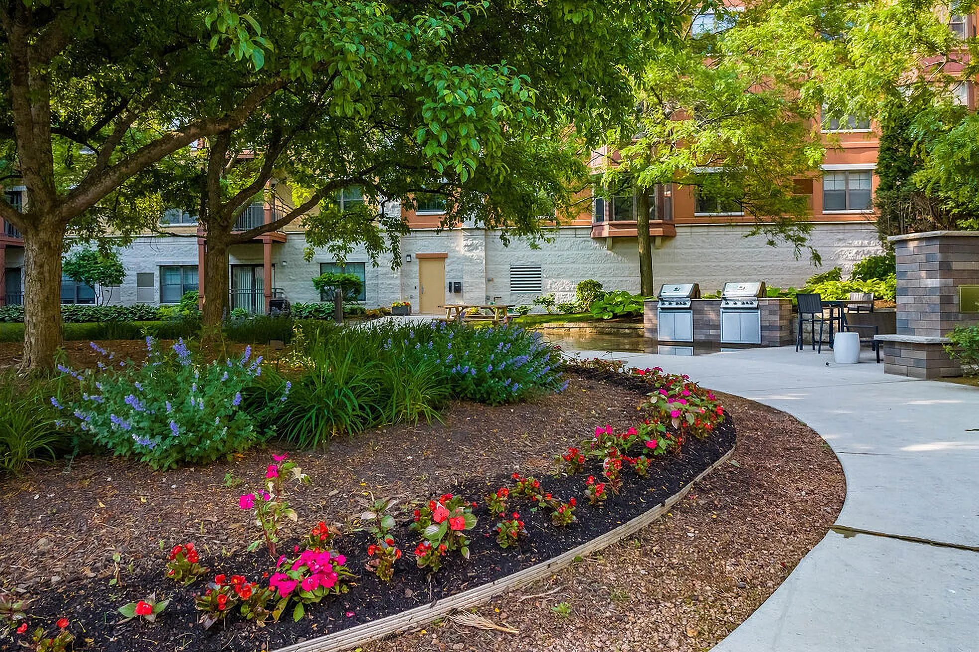Charming garden patio with vibrant flowers, lush greenery, and modern outdoor grills, perfect for entertaining and relaxation in an upscale apartment complex.