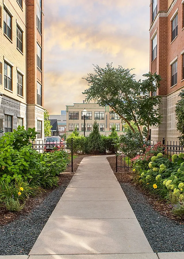Charming apartment courtyard featuring lush landscaping, vibrant flowers, and a welcoming path, nestled between elegant brick buildings.