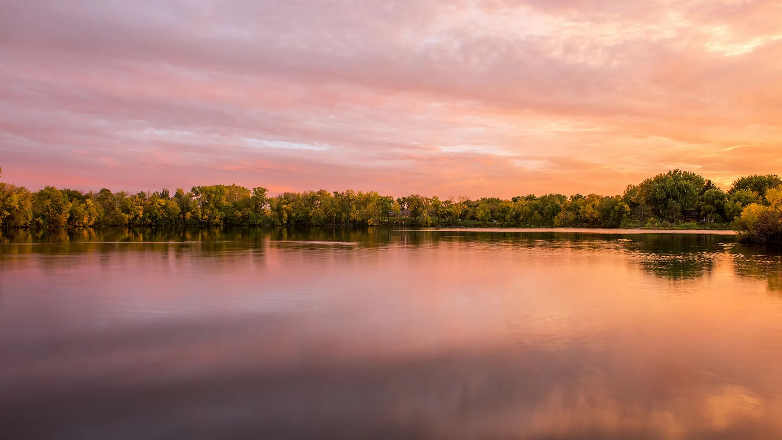Breathtaking lakeside view at sunset with vibrant pink and orange skies reflecting on calm water, surrounded by lush green trees.