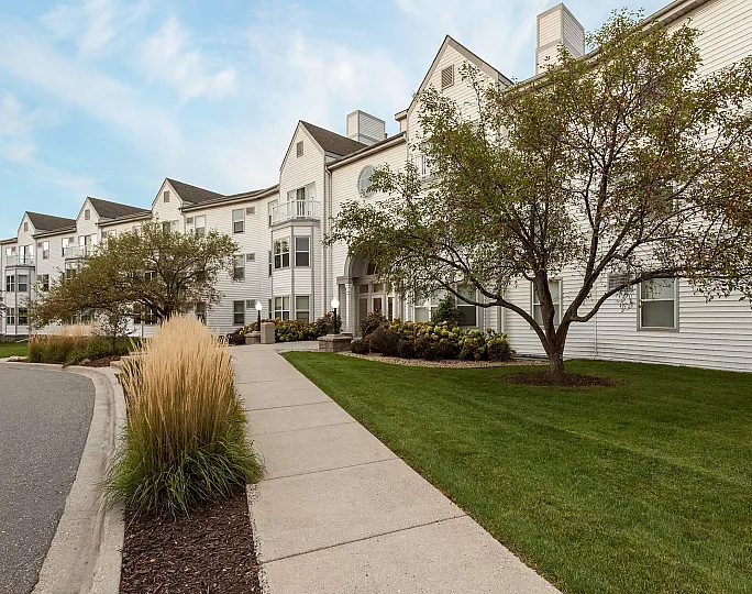 Charming apartment complex with a well-maintained white facade, lush green lawns, and inviting landscaped walkways under a clear blue sky.