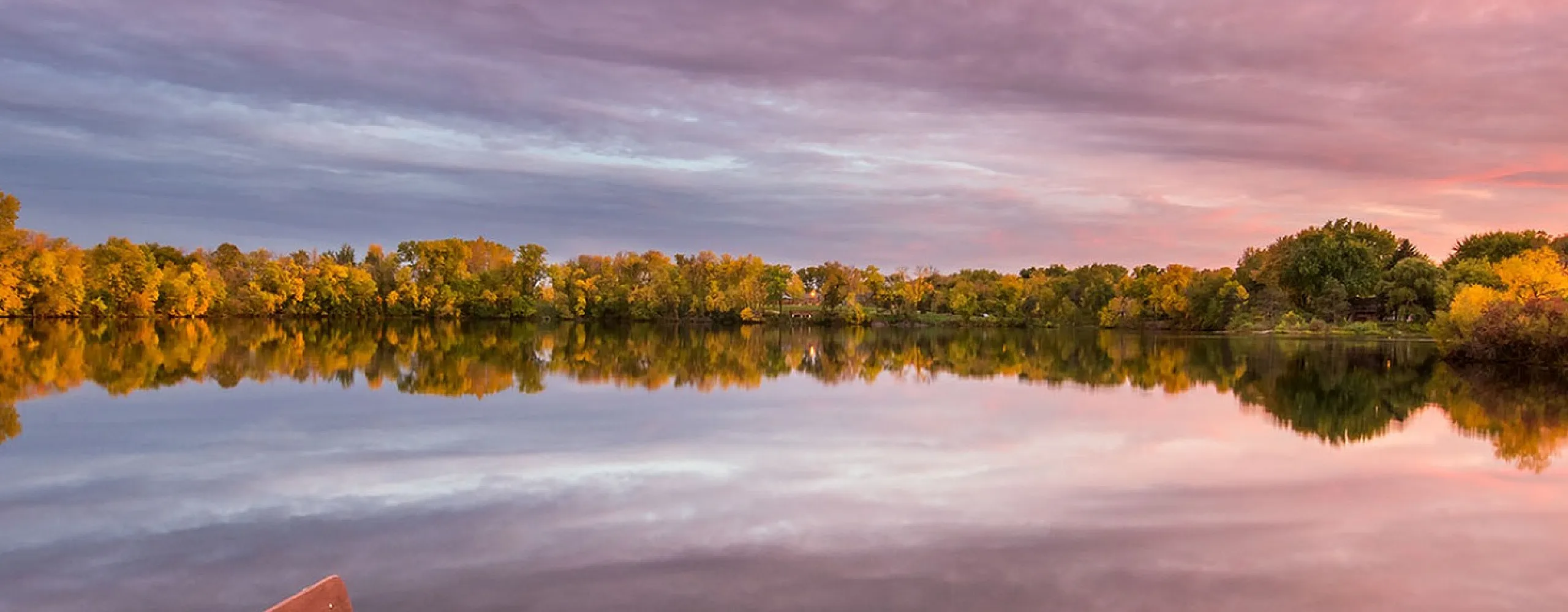 Tranquil lakeside view with a serene bench on a dock, reflecting vibrant sunset hues over lush autumn foliage.
