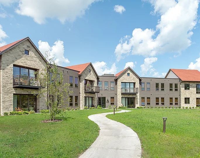 Charming modern apartment building with stone facade and red roofs, nestled in a lush green landscape under a bright blue sky.