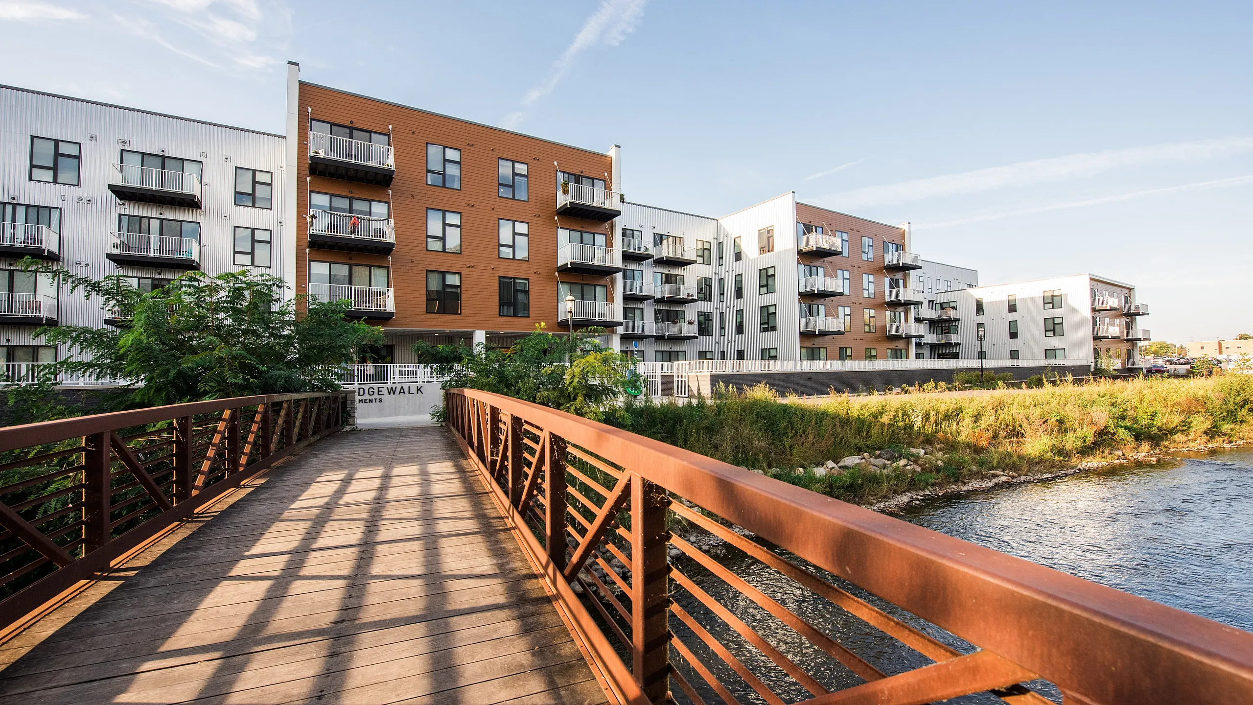 Modern apartment complex with stylish balconies, located by a serene river, connected by a charming wooden bridge.